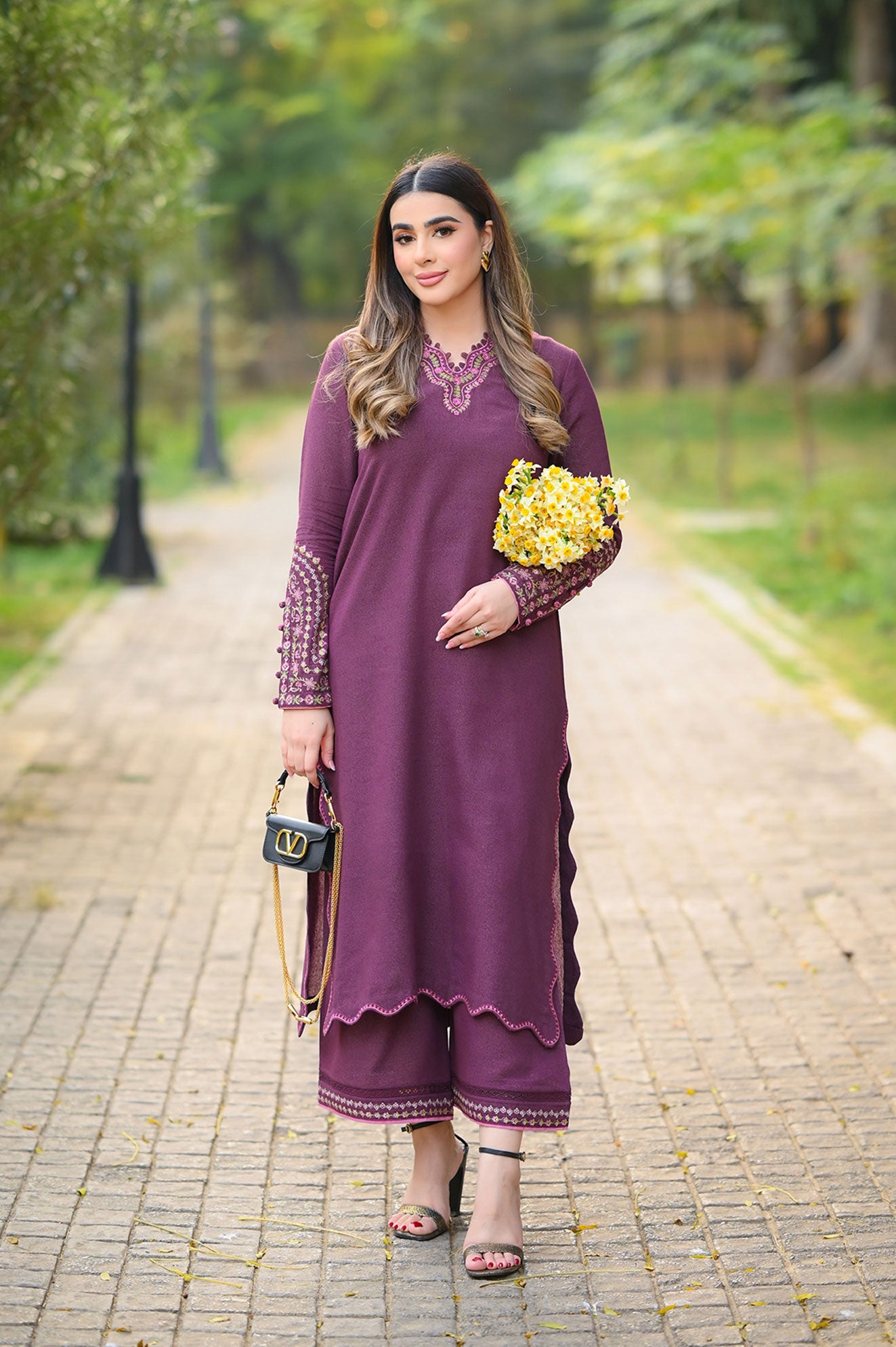 Woman in a purple outfit holding flowers and a handbag on a paved path with greenery.